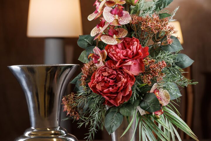 Vista frontale di un mazzo di fiori per cimitero con peonie e orchidee scherziate dai toni autunnali che variano dal rosato al bronzo. La composizione include anche rose e peonie anticate, fogliame verde scuro e accenti di color ruggine. - Mazzo di fiori per cimitero