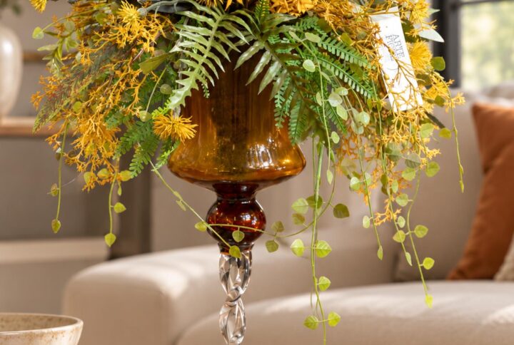 Composizione di fiori artificiali elegante con fiori arancio, dettagli gialli e verde ricadente, in coppa di vetro ambrato con stelo trasparente ritorto.
