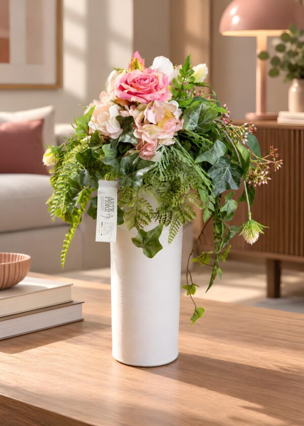 Composizione fiori artificiali in vaso bianco con fiori rosa e avorio, edera variegata e verde ricadente, su un tavolo in soggiorno.