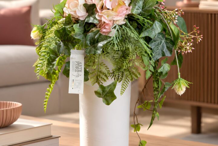 Composizione fiori artificiali in vaso bianco con fiori rosa e avorio, edera variegata e verde ricadente, su un tavolo in soggiorno.