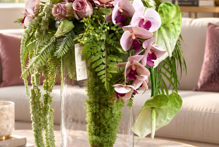 Composizione floreale con verde ricadente e orchidee rosa in vaso cilindrico trasparente, con licheni artificiali e fondo fucsia.