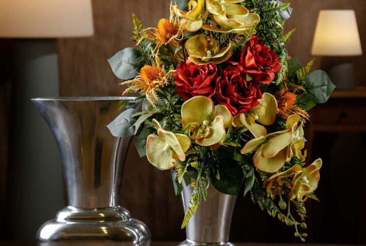 Vista di un mazzo floreale in primo piano con fiori artificiali arancioni e gialli in vaso argentato – Un’elegante composizione floreale con orchidee gialle, rose rosse e dettagli in verde per loculi cimiteriali.