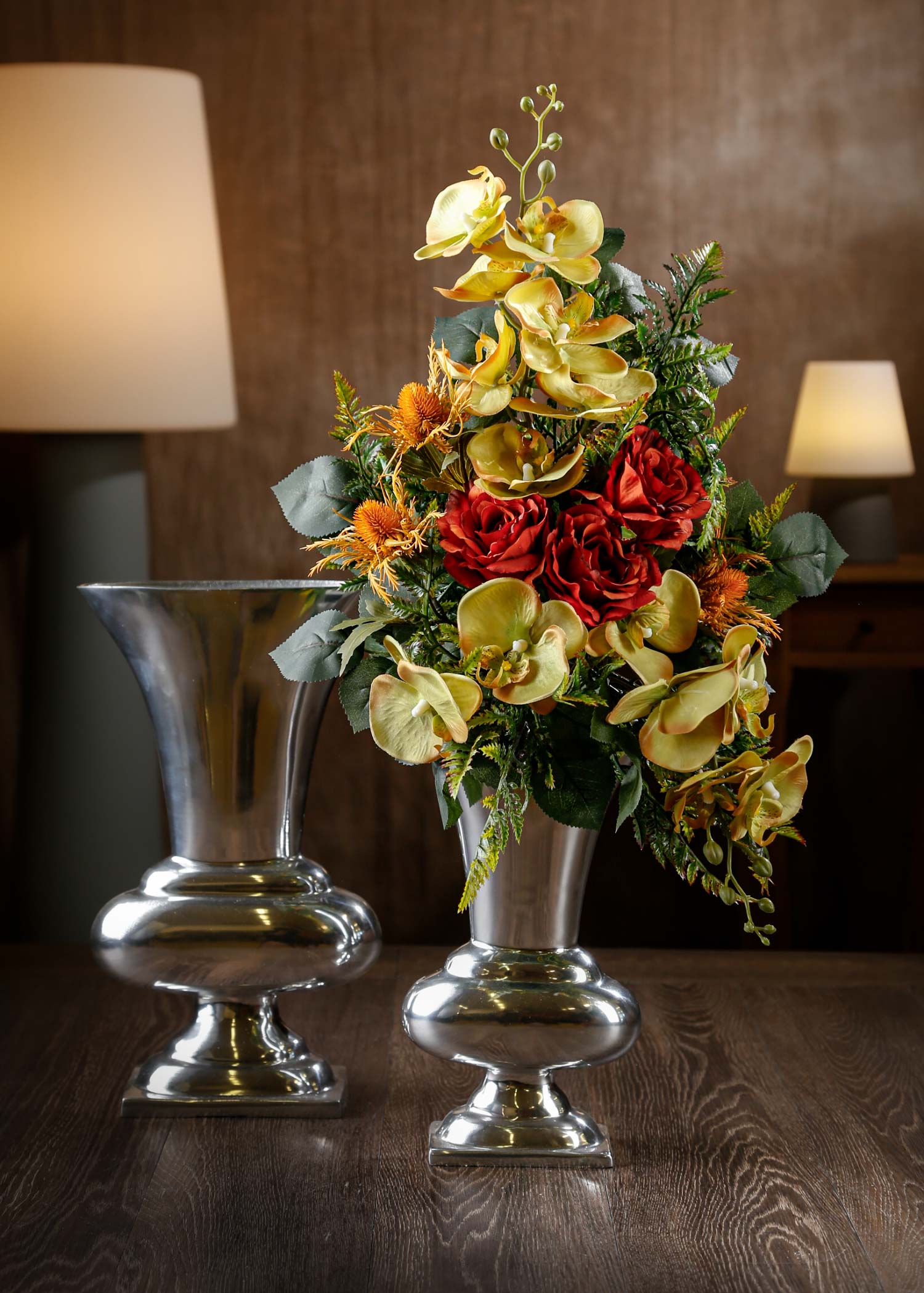 Vista di un mazzo floreale in primo piano con fiori artificiali arancioni e gialli in vaso argentato – Un’elegante composizione floreale con orchidee gialle, rose rosse e dettagli in verde per loculi cimiteriali.