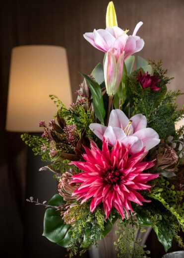 Dettaglio ravvicinato della composizione di fiori finti per cimitero con gigli rosa e dalie fucsia circondati da fogliame verde.