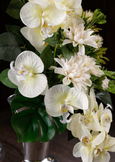 Composizione floreale con orchidee e dalie bianche in un bouquet di fiori finti per cimitero, posizionato su un vaso argentato